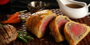 A golden-brown Beef Wellington sliced open to reveal its tender, pink center, served on a rustic wooden board with rosemary garnish and a side of gravy.