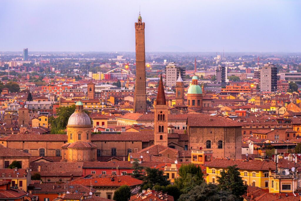 A panoramic view of Bologna at sunset, featuring the iconic Asinelli Tower rising above terracotta rooftops and historic domed churches in the city center.