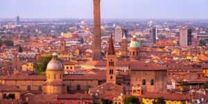 A panoramic view of Bologna at sunset, featuring the iconic Asinelli Tower rising above terracotta rooftops and historic domed churches in the city center.