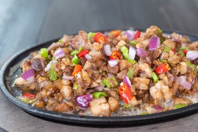 A sizzling plate of Filipino pork sisig topped with red onions and chilies, served on a hot iron skillet.