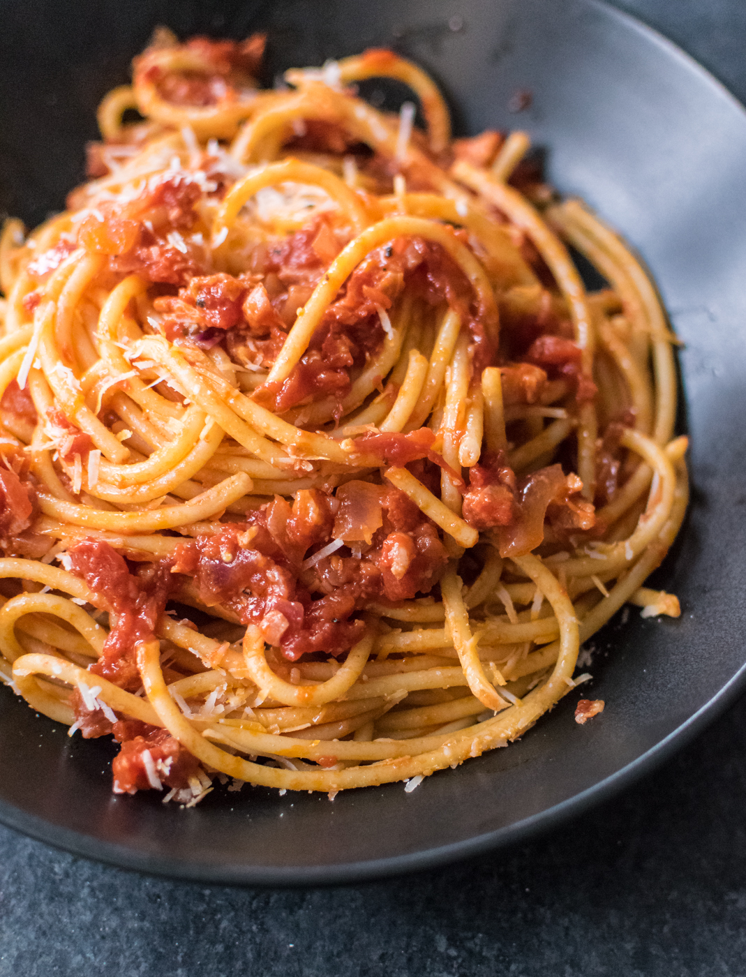 A close-up of bucatini pasta coated in a rich tomato and guanciale sauce, topped with grated pecorino cheese, served in a dark ceramic bowl.