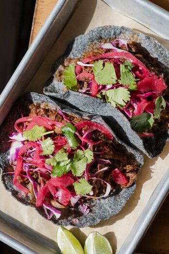 Two blue corn tortillas filled with juicy short rib barbacoa, red onions, and cilantro, served on a metal tray with lime wedges.