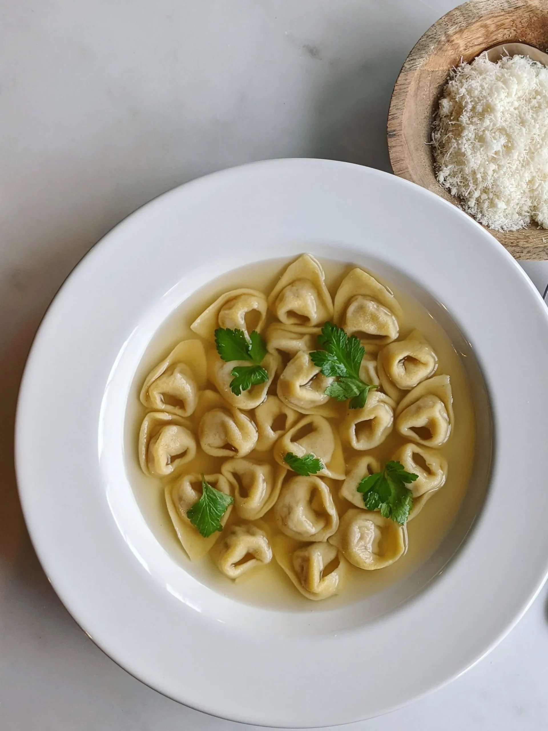 A white bowl of tortellini in clear broth, garnished with parsley leaves, served alongside a wooden bowl of grated Parmigiano Reggiano and a silver spoon.