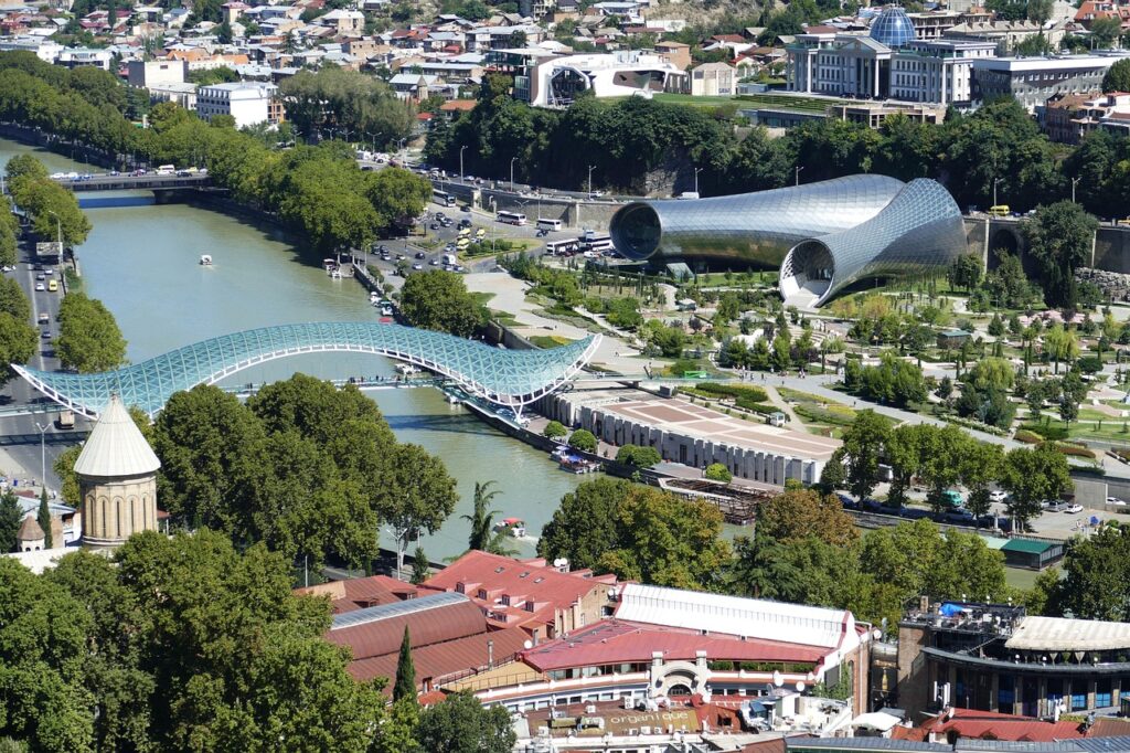 The Bridge of Peace stretching over the Kura River in Tbilisi, Georgia, with Rike Park and the modern tubular concert hall in the background — a symbolic view of East meets West.