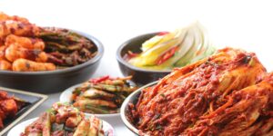 A colorful assortment of traditional Korean Kimchi dishes, including napa cabbage kimchi, green onion kimchi, radish kimchi, and young napa cabbage, all arranged in bowls and plates on a bright surface.