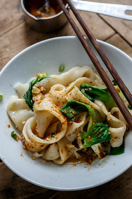 A bowl of wide hand-pulled noodles topped with chili oil and bok choy, served with chopsticks on a rustic wooden table.