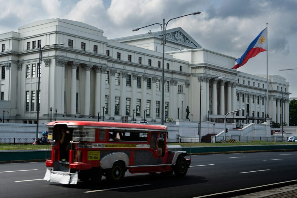A red and silver jeepney drives past the National Museum of Fine Arts in Manila, with the Philippine flag waving under a cloudy sky — a powerful blend of history, identity, and everyday life in the capital.