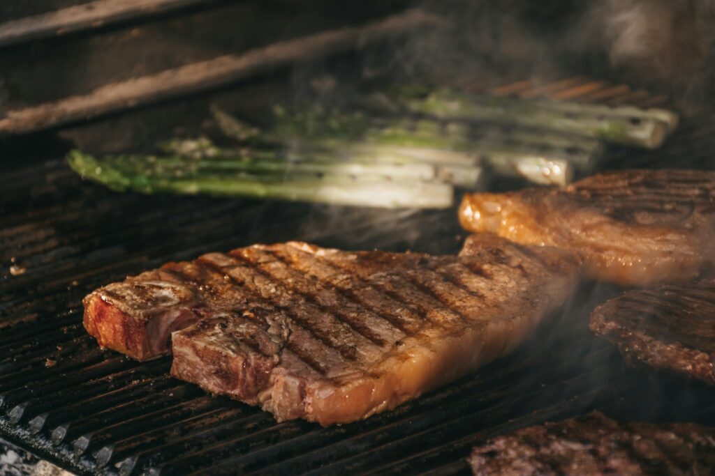 Thick Florentine T-bone steak grilling over open flames, with charred grill marks and green asparagus cooking alongside.