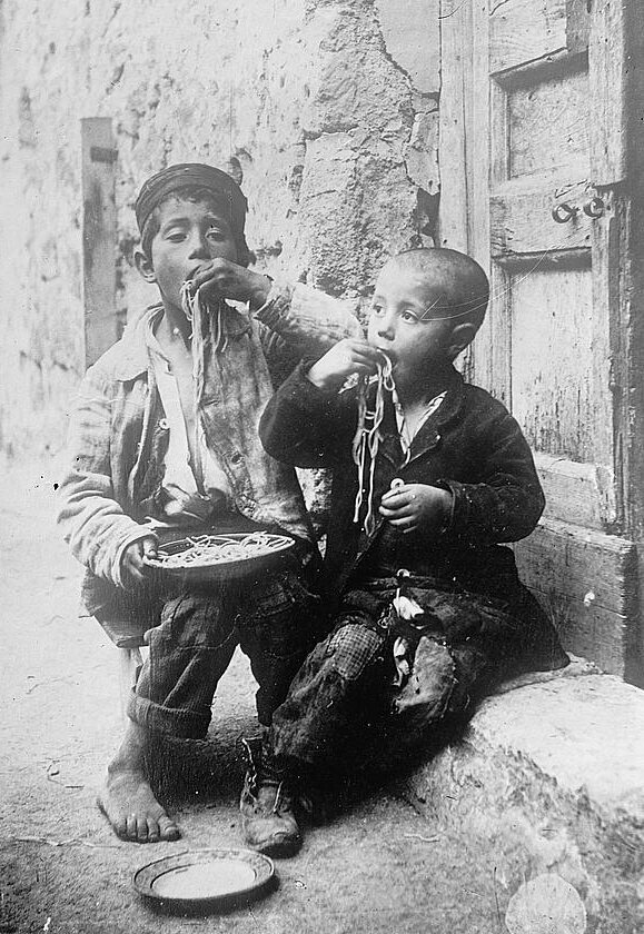 Late‑19th‑century street children eating spaghetti by hand on a street corner
