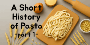 Freshly made fettuccine on a wooden board with eggs, semolina, wheat stalks, and a rolling pin – title overlay reads ‘A Short History of Pasta – Part 1