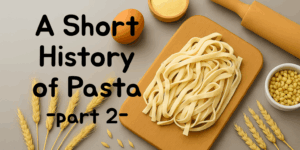 Freshly made fettuccine on a wooden board with eggs, semolina, wheat stalks, and a rolling pin – title overlay reads ‘A Short History of Pasta – Part 2