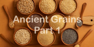 Assorted ancient grains including farro, chia, millet, and sorghum displayed in round wooden bowls on a wooden cutting board, with wooden spoons holding small portions, set against a warm-toned background.