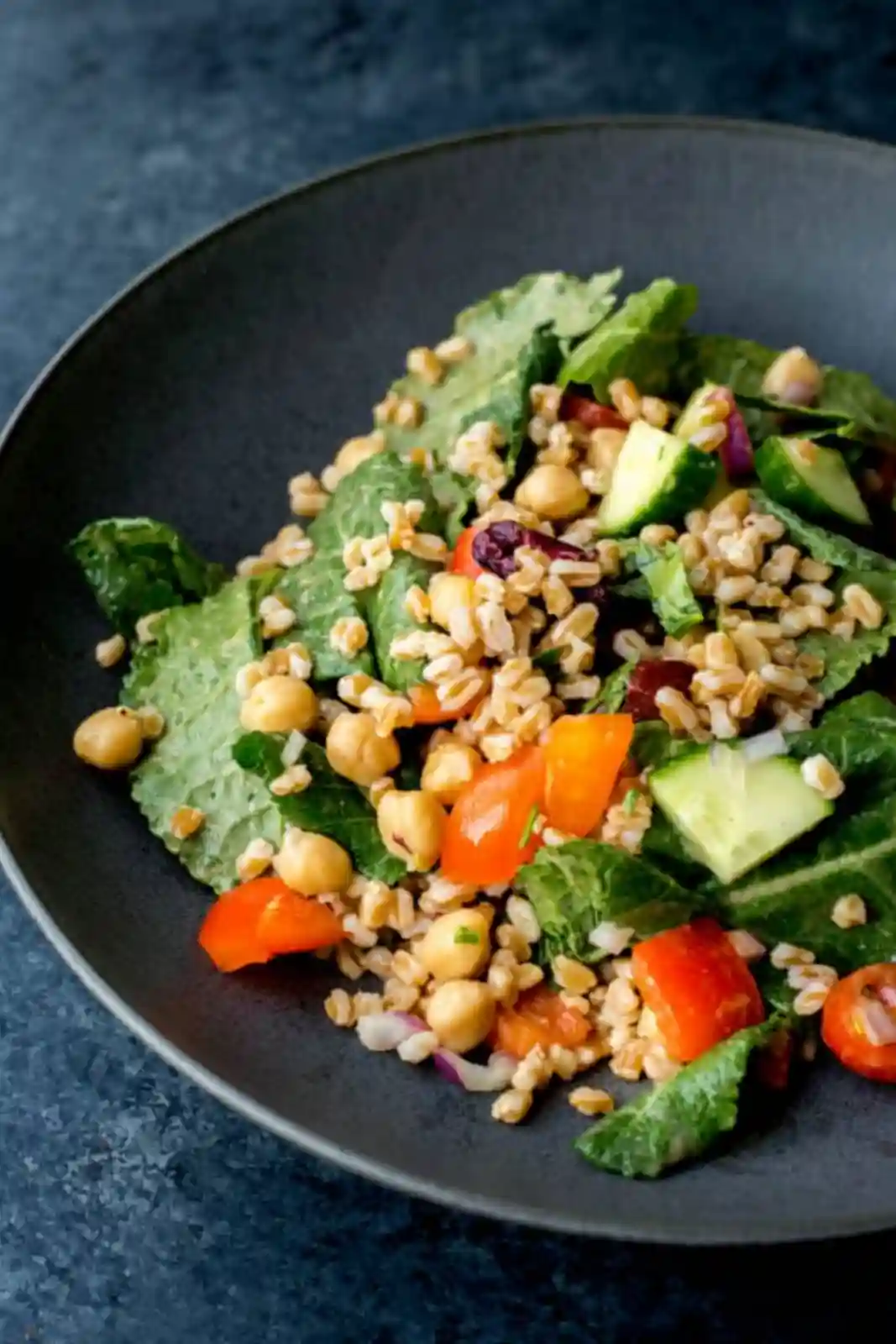 A bowl of ancient grain salad with farro, chickpeas, chopped vegetables, and fresh greens on a dark plate.