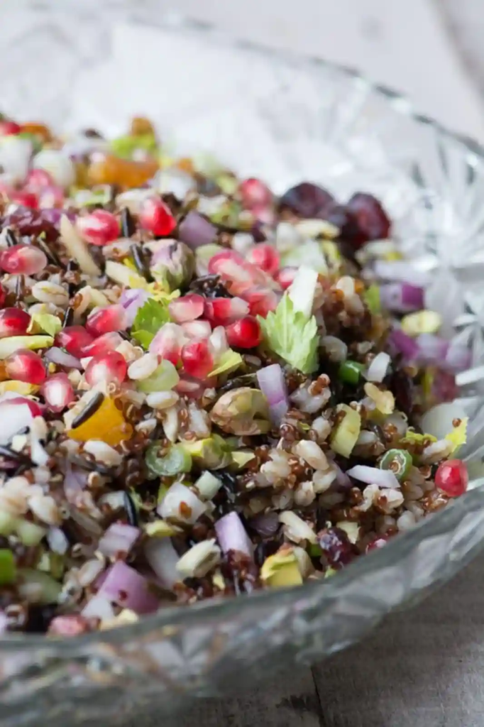 A colorful wild rice and quinoa salad mixed with pomegranate seeds, pistachios, and chopped vegetables in a glass bowl.