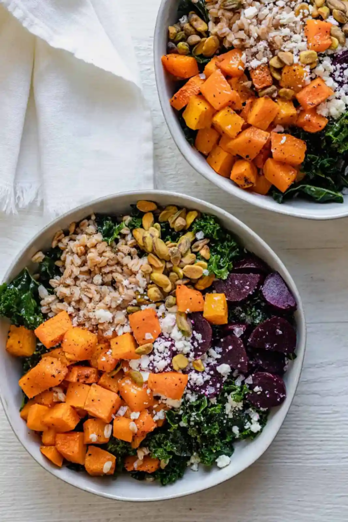 Colorful grain bowls topped with farro, roasted sweet potatoes, kale, beets, pistachios, and crumbled cheese.