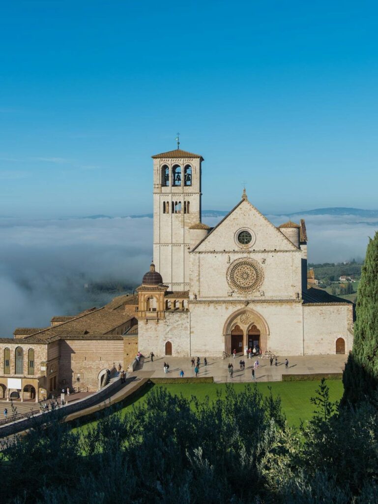 Front view of the Basilica of Saint Francis in Assisi, Umbria, with visitors and clear blue sky, featured in Stanley Tucci Searching for Italy