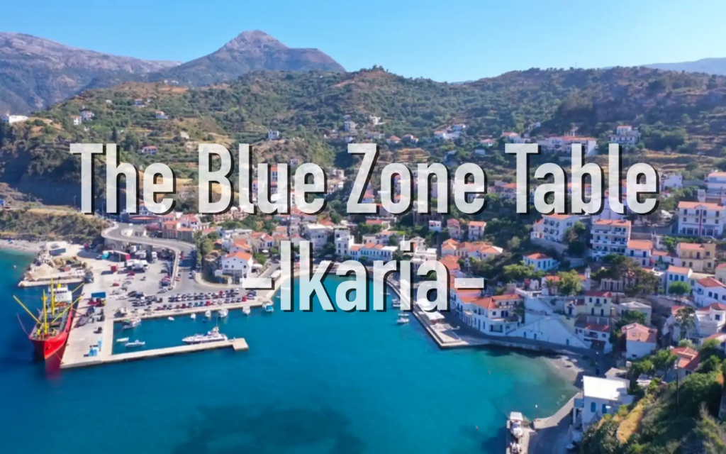 A scenic aerial view of a coastal village in Ikaria, Greece, with red-roofed houses, mountains in the background, and turquoise water in the harbor.
