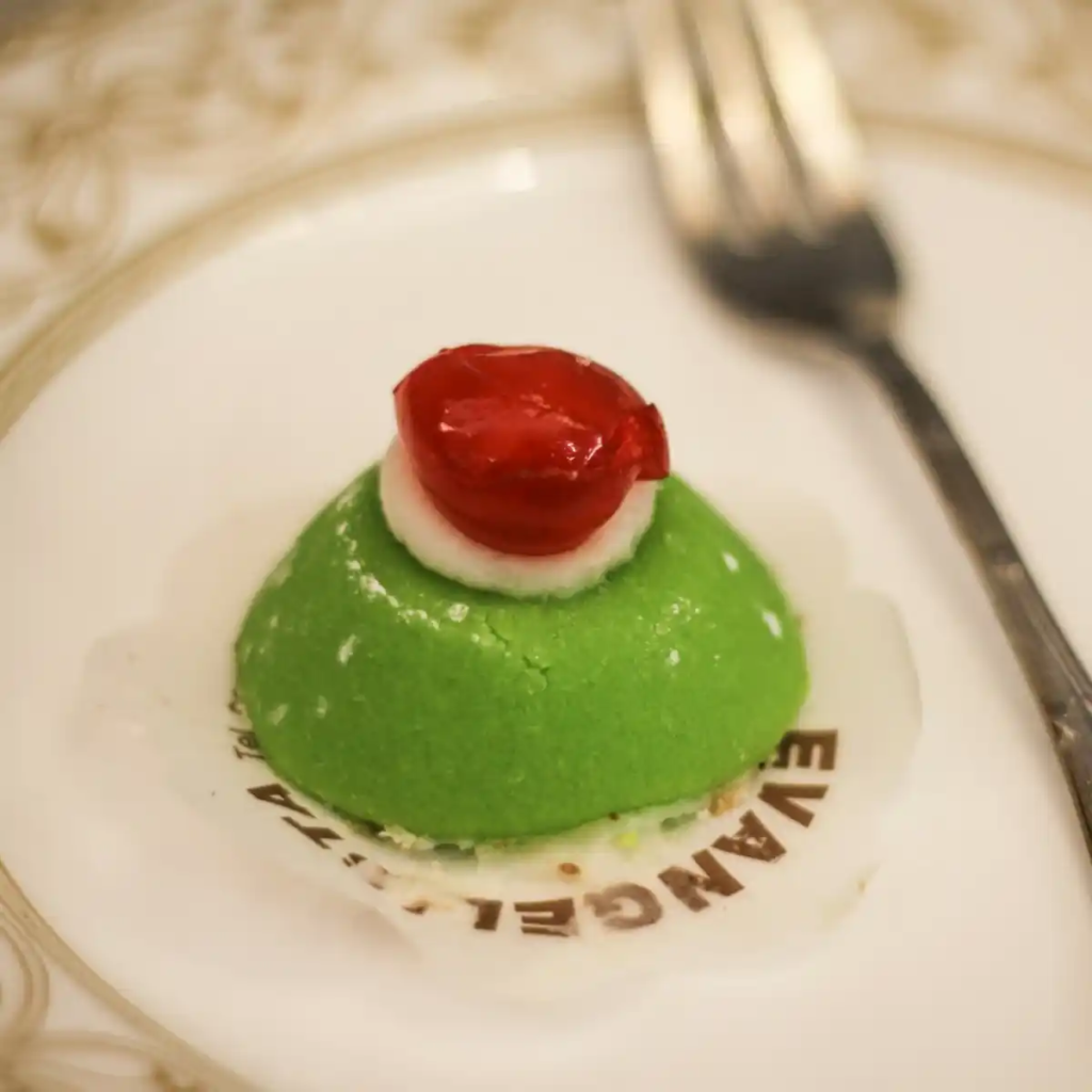 A brightly colored Sicilian cassata topped with a red candied cherry, wrapped in green marzipan on a white plate.