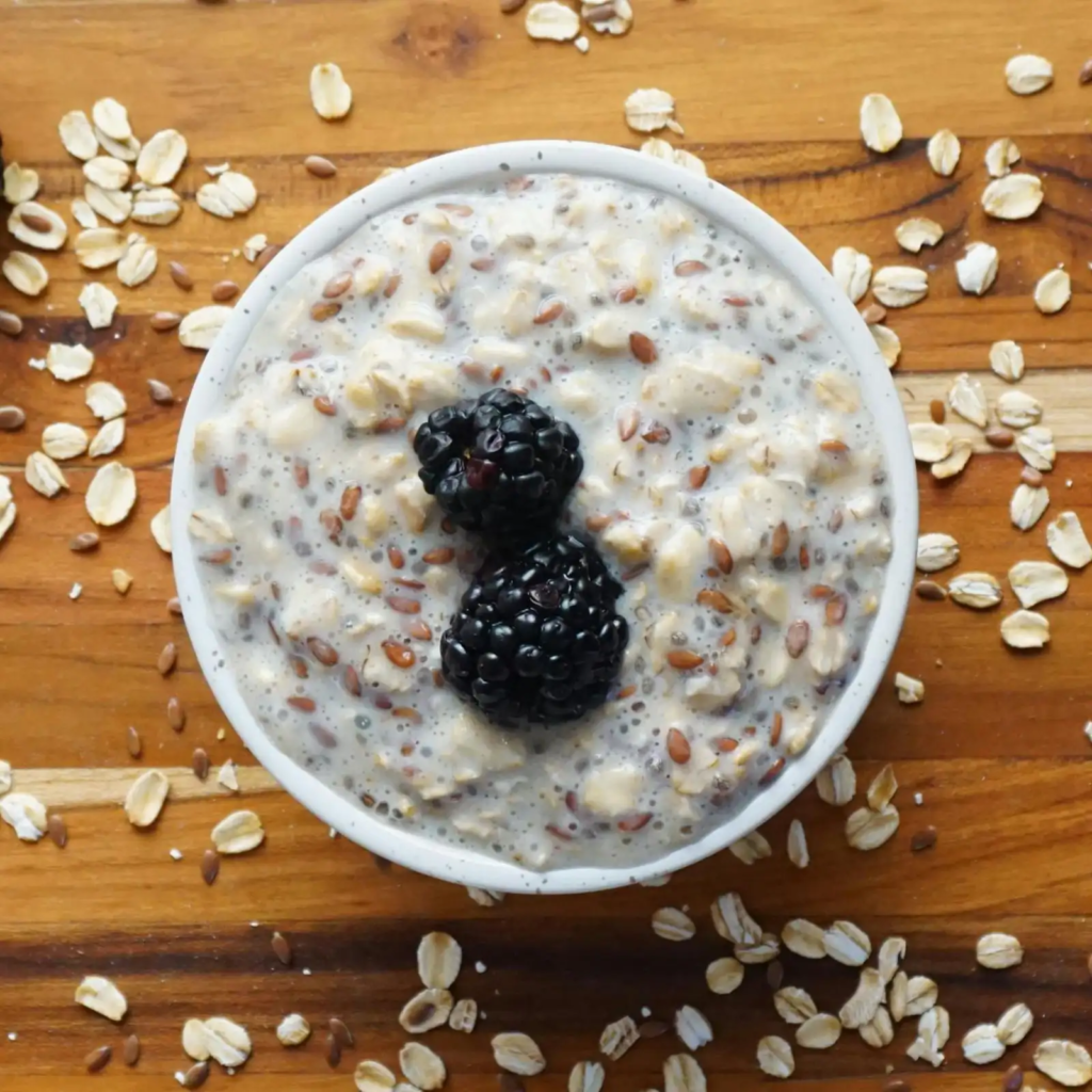 A white bowl filled with creamy overnight chia oatmeal topped with two blackberries, surrounded by scattered oats on a wooden surface.