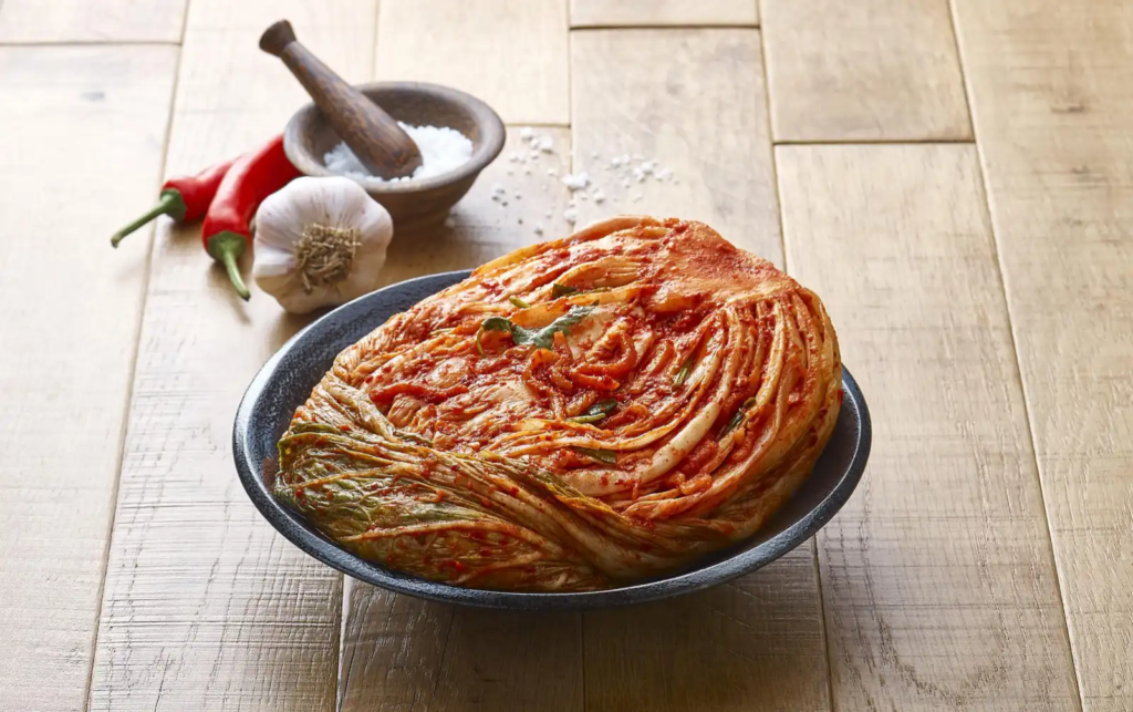 Whole napa cabbage Kimchi in a bowl with garlic, chili, and salt in the background – a staple Korean fermented food
