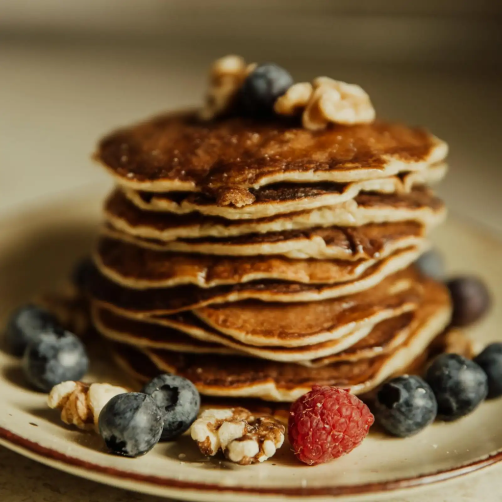 A tall stack of golden-brown protein pancakes topped with fresh blueberries, raspberries, and walnut pieces on a ceramic plate.
