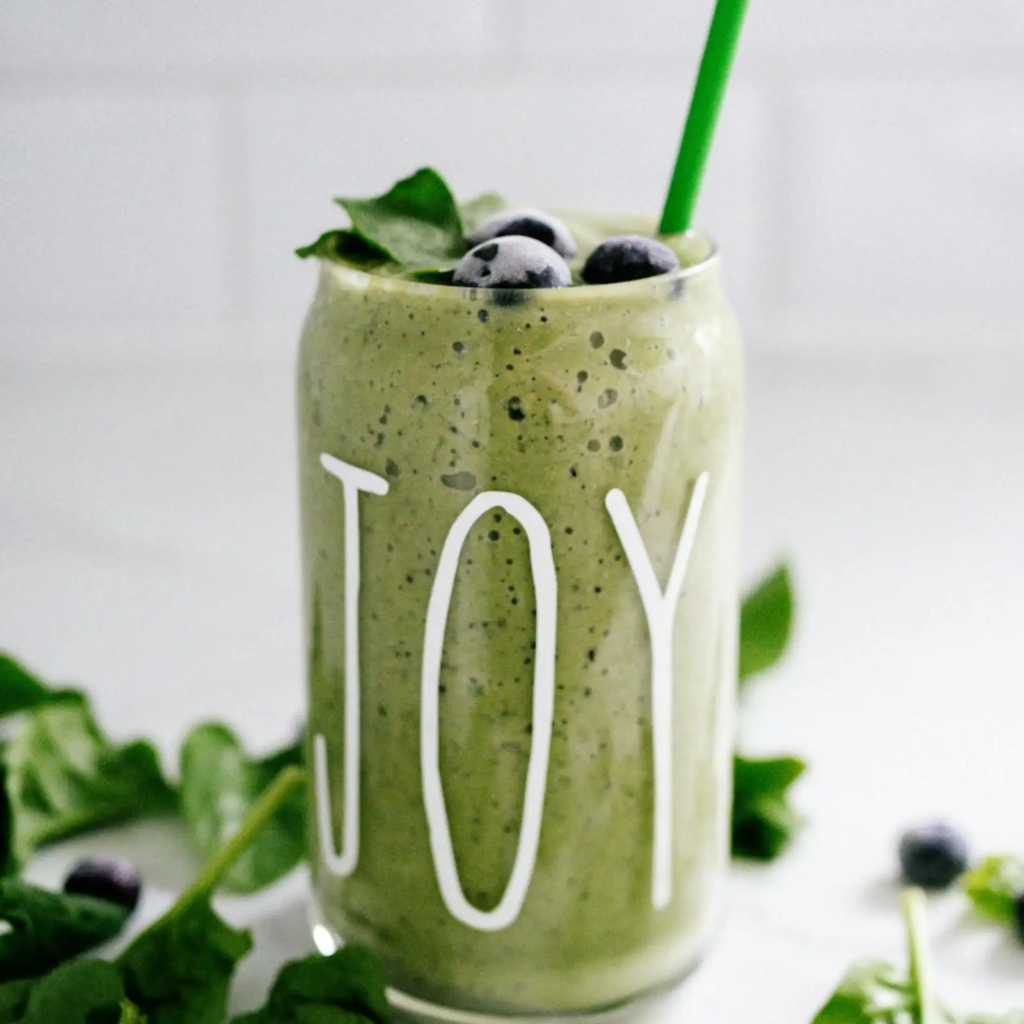 A tall glass labeled “JOY” filled with green smoothie speckled with chia seeds, topped with blueberries and spinach leaves.