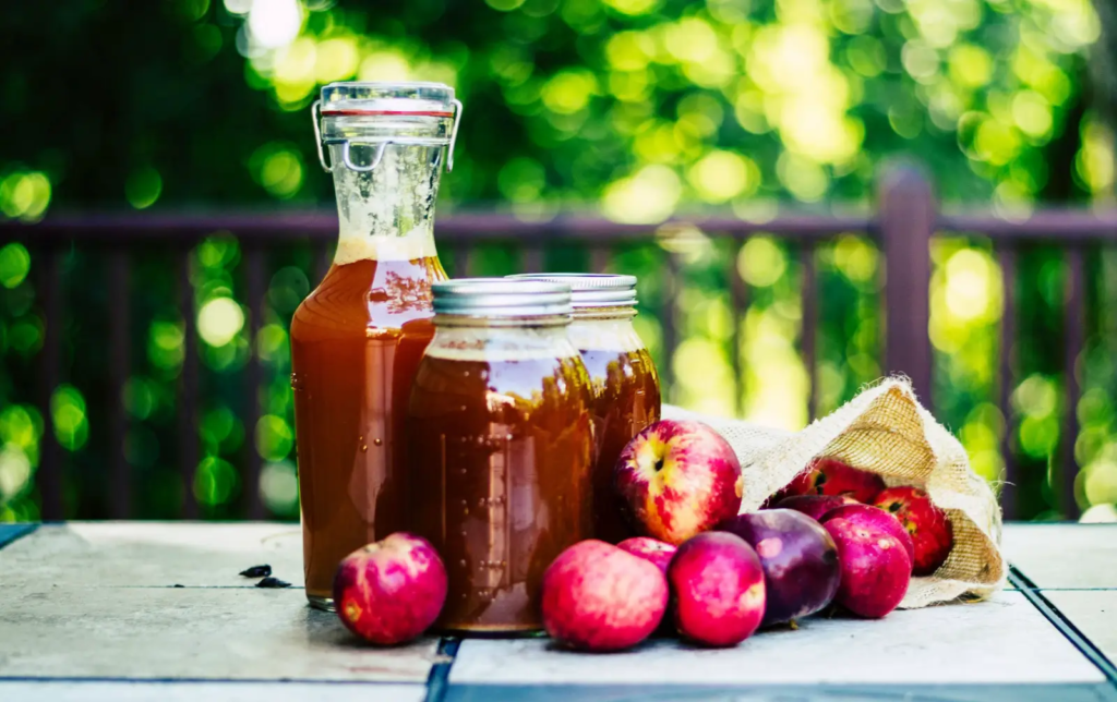 Bottles and mason jars of fresh-pressed apple cider on an outdoor table, with red apples and a green orchard backdrop.