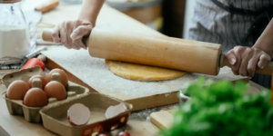 Hands rolling fresh pasta dough with a wooden rolling pin on a floured surface, surrounded by eggs, flour, and fresh vegetables.