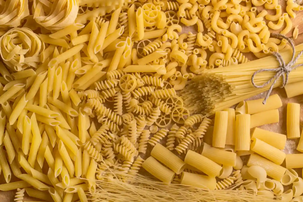 Assorted shapes of dried pasta, including penne, fusilli, rigatoni, and spaghetti, arranged on a warm-toned surface.