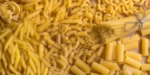 Assorted shapes of dried pasta, including penne, fusilli, rigatoni, and spaghetti, arranged on a warm-toned surface.