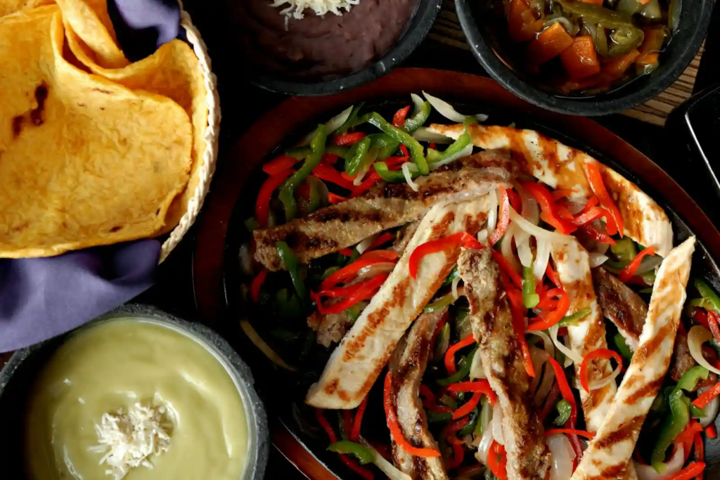 Close-up of chicken and beef fajitas with grilled peppers and onions served with tortillas and sauces.
