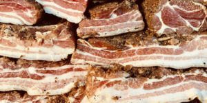 Close-up of cured pork belly slices stacked together, showing layers of meat and fat with seasoning.