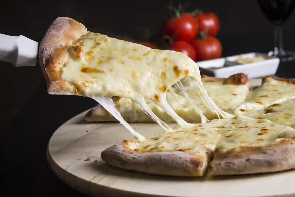 Close-up cheese pizza slice being lifted with long stretchy mozzarella strands on a wooden board; tomatoes softly blurred in the background.