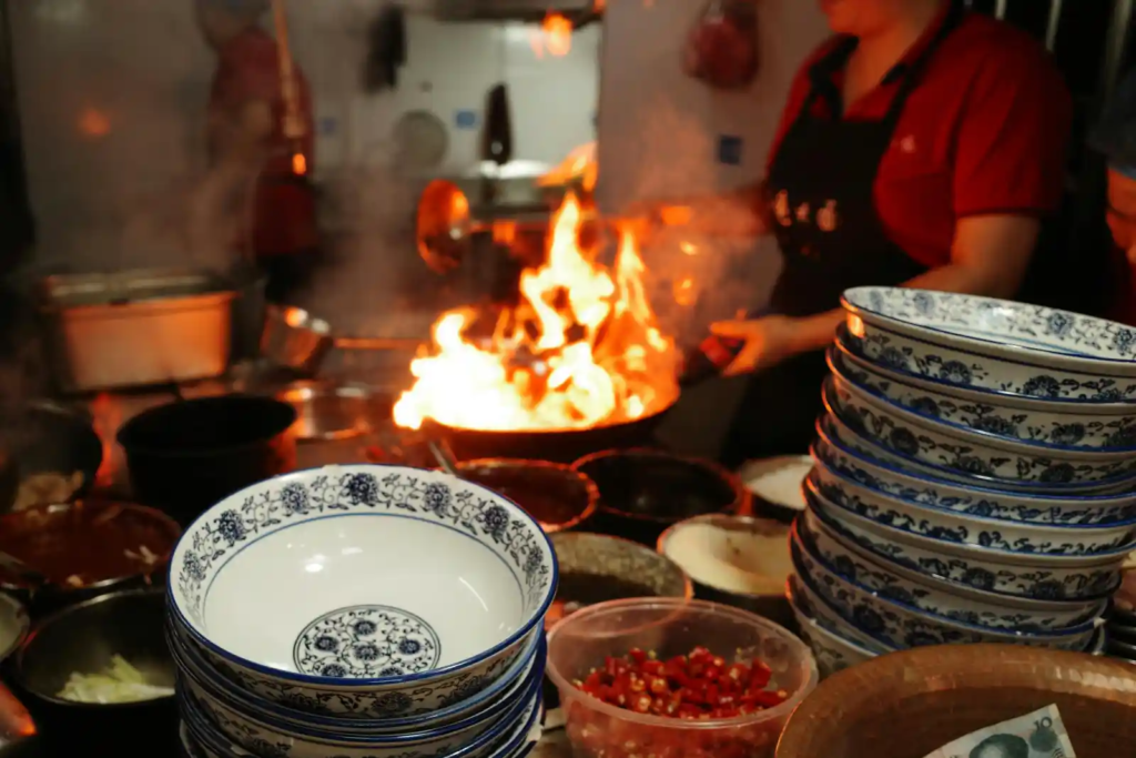 Chef stir-frying food over high flames in a busy restaurant kitchen.
