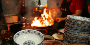 Chef stir-frying food over high flames in a busy restaurant kitchen.