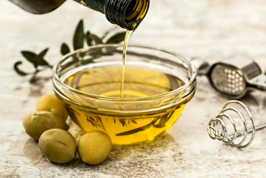 Golden olive oil being poured into a glass bowl, with fresh green olives and a whisk nearby.