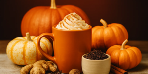 Pumpkin spice latte in an orange mug topped with whipped cream and cinnamon, surrounded by pumpkins, ginger, nutmeg, cloves, and cinnamon sticks.