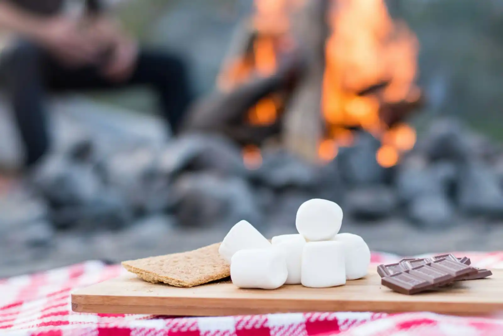 Graham crackers, marshmallows, and chocolate stacked on a wooden board in front of a campfire.