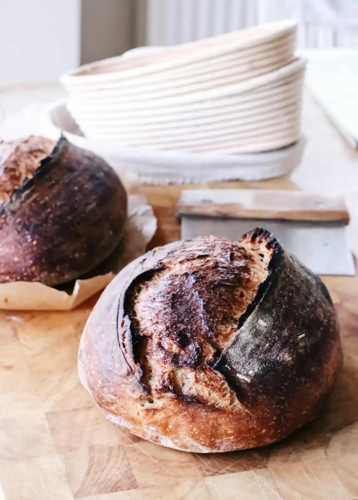 Two deeply caramelized sourdough loaves resting on a wooden surface, with a bench scraper and stacked proofing baskets in the background, all softly lit by natural light.
