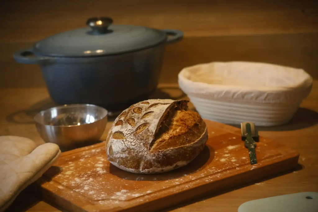 A freshly baked sourdough loaf on a floured wooden board, surrounded by essential baking tools including a proofing basket, a Dutch oven, a lame, and a mixing bowl—set in a warmly lit kitchen.