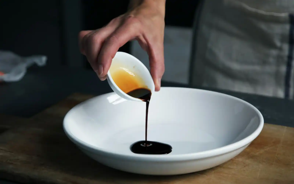 A close-up of a hand pouring dark soy sauce from a small white bowl into a wide white plate on a wooden surface.