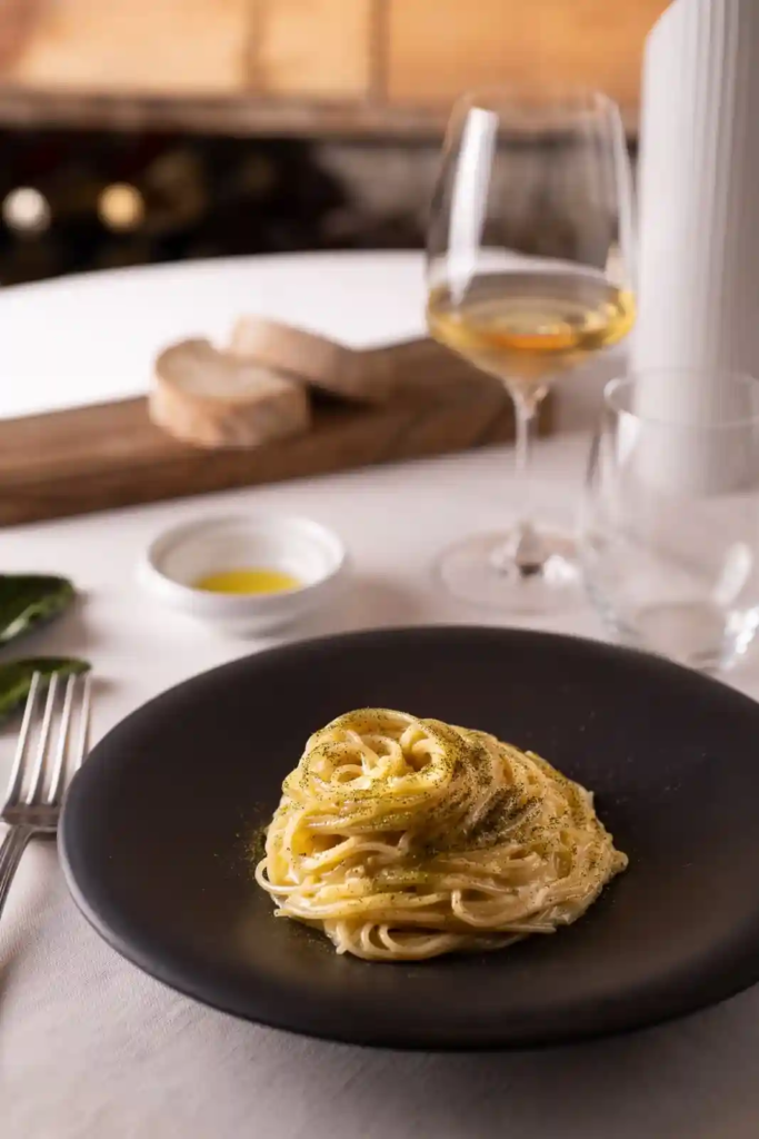  Plate of spaghettini pasta coated in a creamy lemon sauce, served on a black plate with wine in the background.