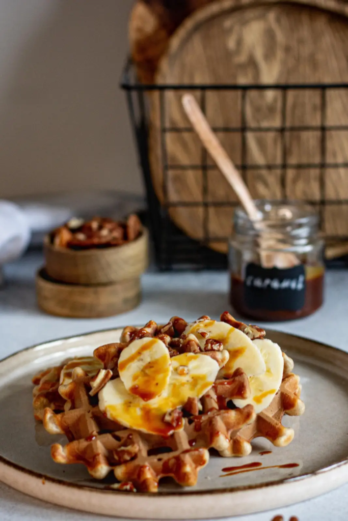 A Belgian waffle topped with banana slices, chopped nuts, and caramel drizzle.