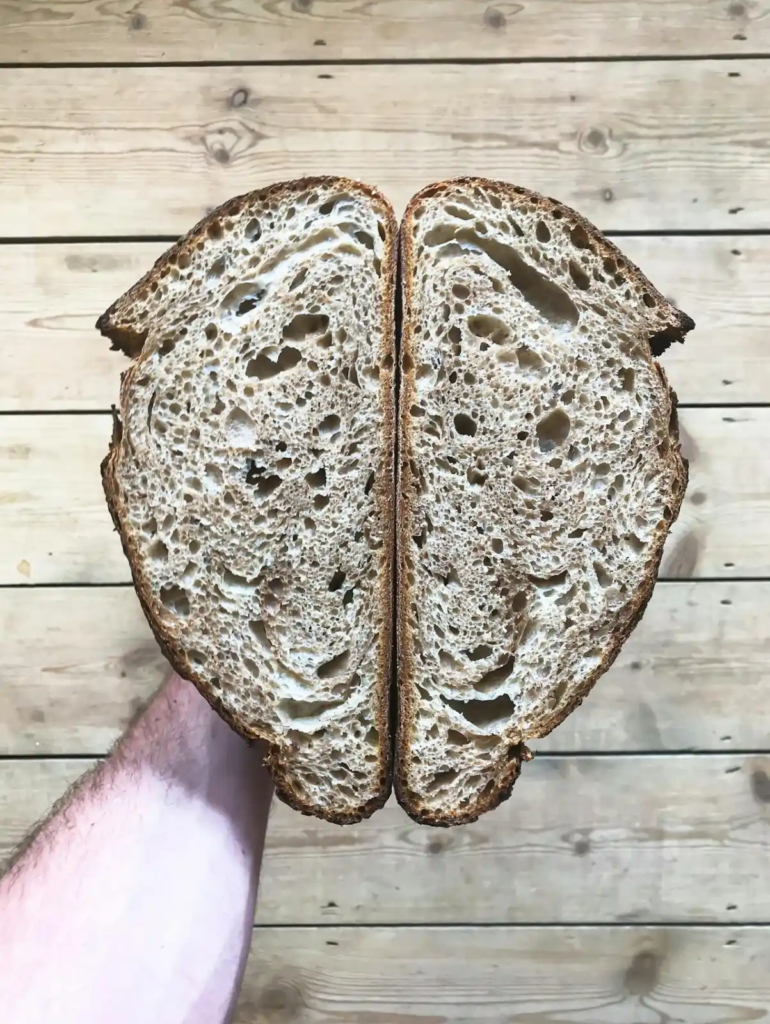 A hand holding two halves of a sliced sourdough loaf, showing the airy, open crumb structure against a rustic wooden background.