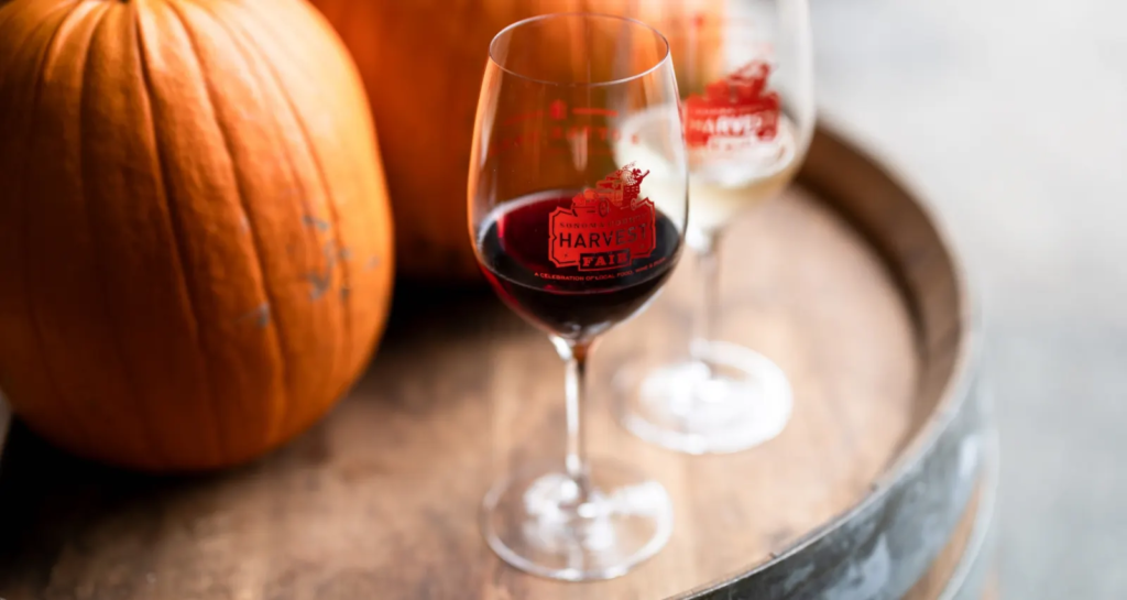 Harvest Fair–branded wine glass with red wine on a wooden barrel; pumpkins and a second glass softly blurred in the background.