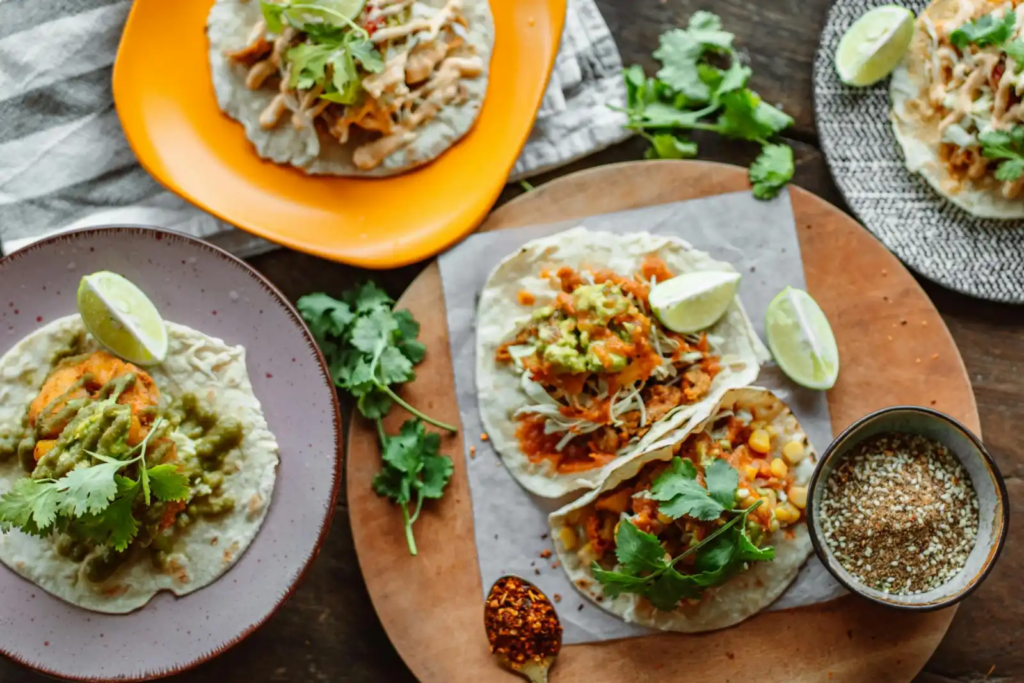 Assorted tacos on colorful plates topped with guacamole, cilantro, and lime wedges, served with spices on a rustic wooden table.