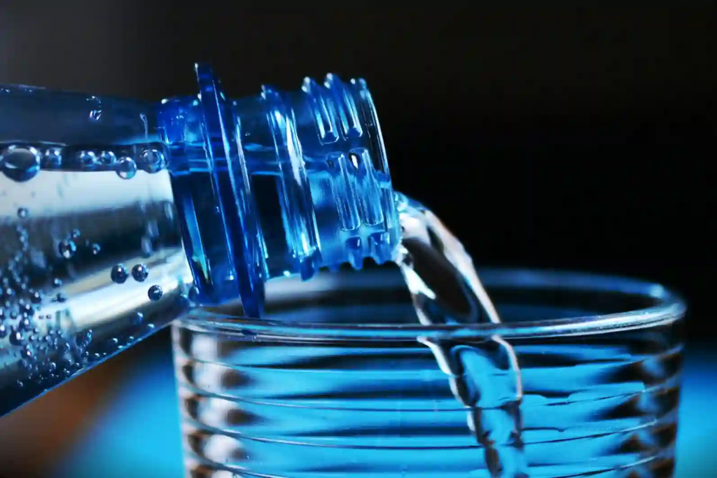 Close-up of water being poured from a blue plastic bottle into a ridged glass, with crisp blue lighting and tiny droplets on the bottle.