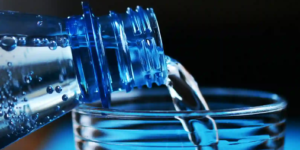 Close-up of water being poured from a blue plastic bottle into a ridged glass, with crisp blue lighting and tiny droplets on the bottle.